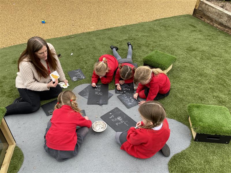 5 children in school red school jumpers lay on the floor with a teacher practicing phonics on pentagon plays scribble boards