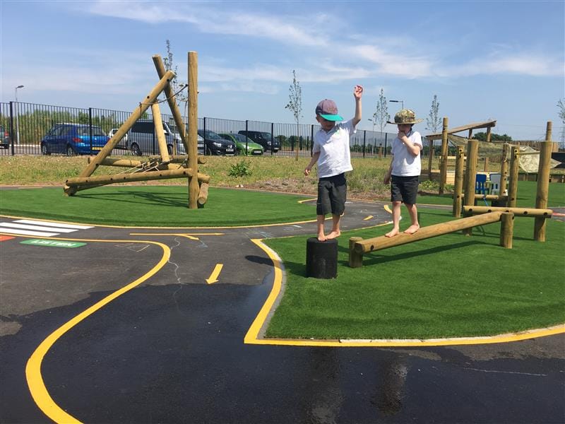 Two children in shorts, white polos and sun hats playing in the school playground. One is balancing on a black stepping log and the other is balancing on an inclined balance beam