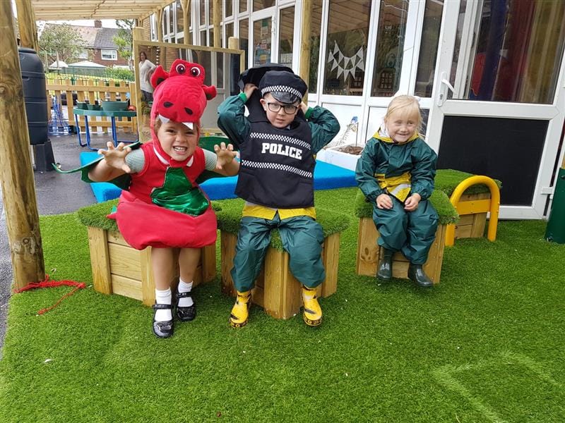 Three children are sat on artificial grass topped seats as they pose to the camera. 2 children are dressed up in a different costume, with one being a dinosaur and the other being a police officer. The final child is wearing their coat and wellies.