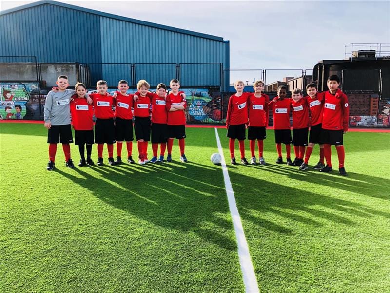 Two football teams are stood on a football pitch as they stand side-by-side with a football between them. The children are looking at the camera and are smiling.