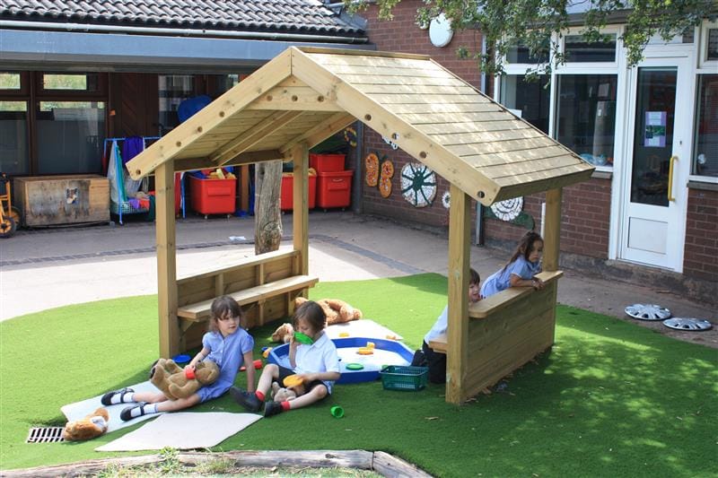four children sit underneath a playground playhouses with benches and have a variety of toys and books to play with and read