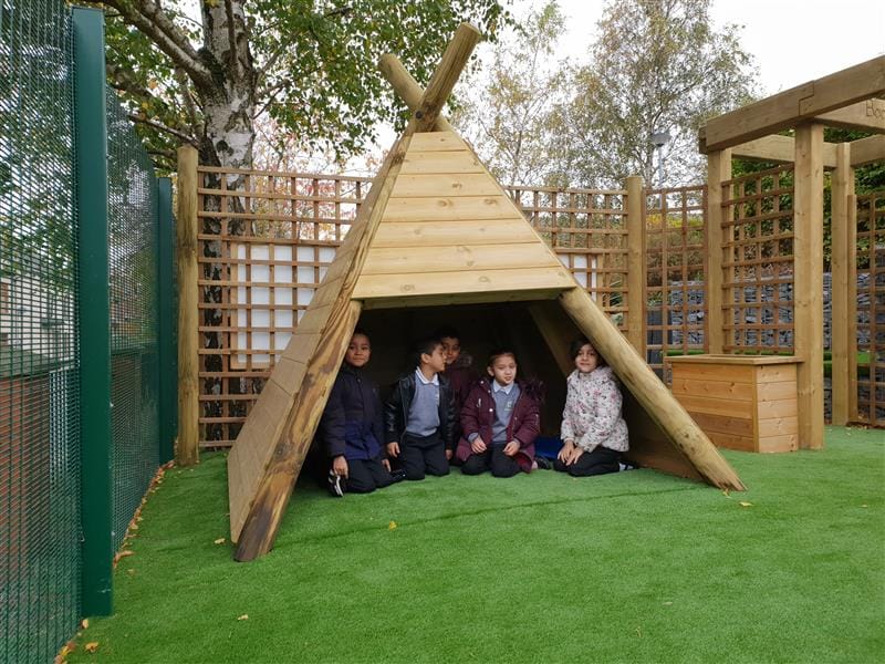 five children sit sheltered under a wigwam on the green artificial grass, they all wear winter coats