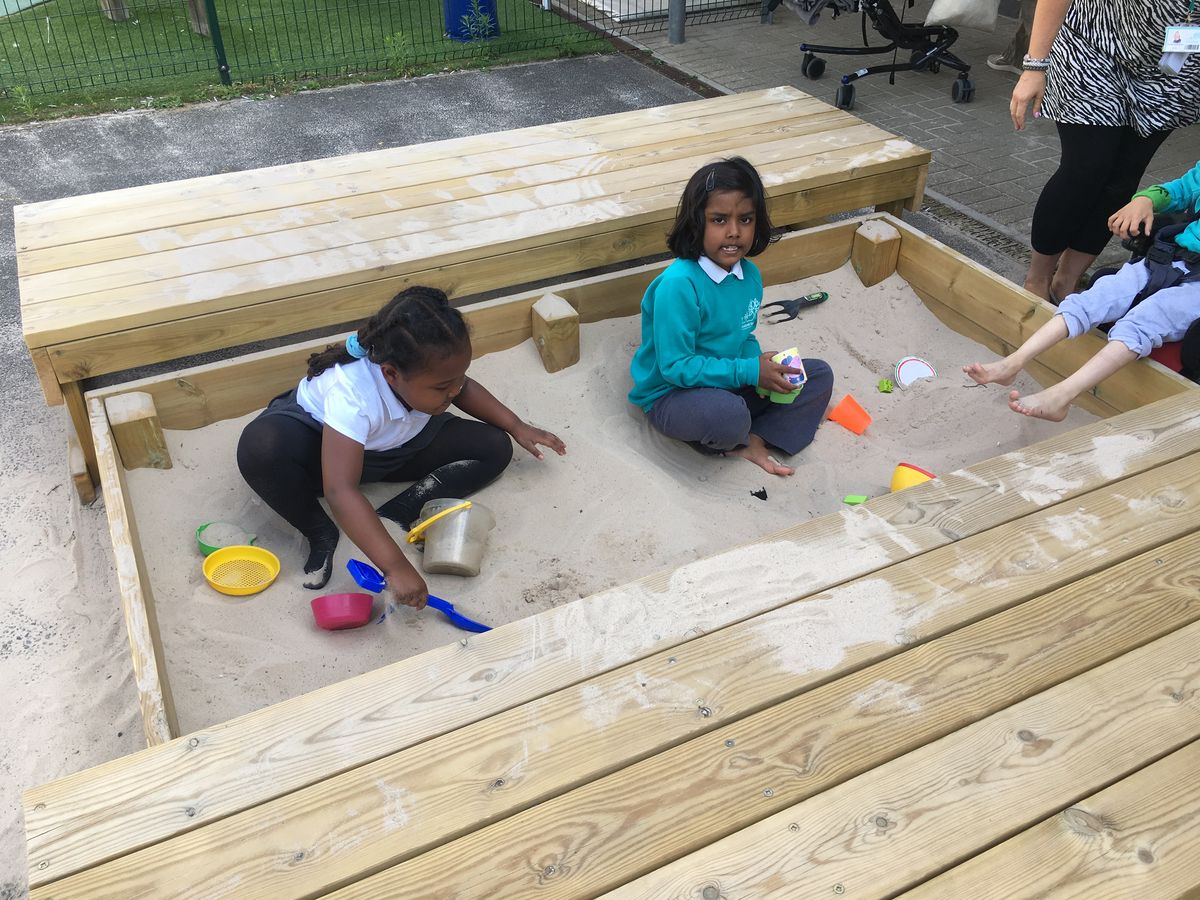 two chldren in turquoise uniform sit in the sand pit and play with sand and look up at the camera