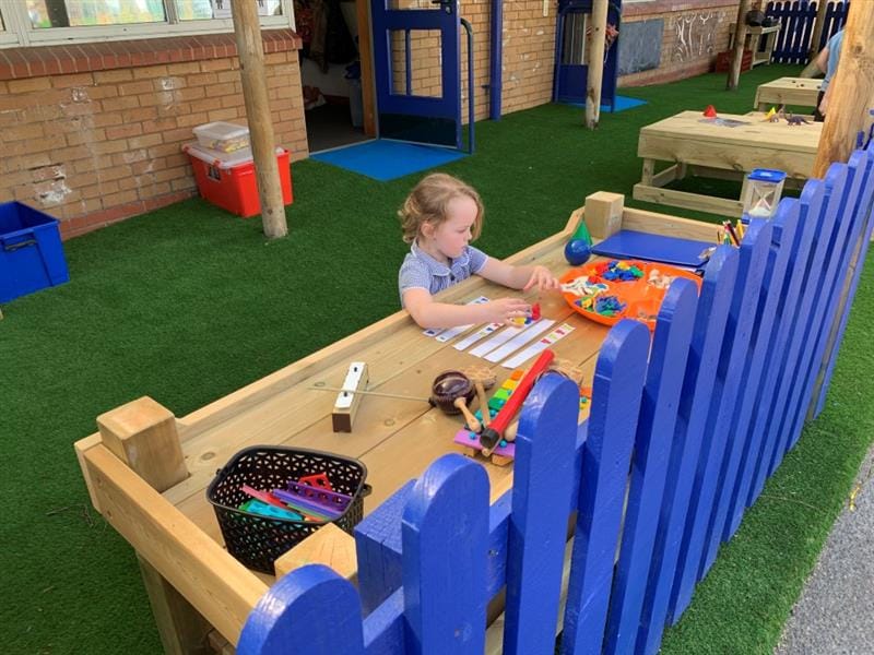 a little girl in a blue school dress sits and draws at a construction table with artiicial grass surfacing below