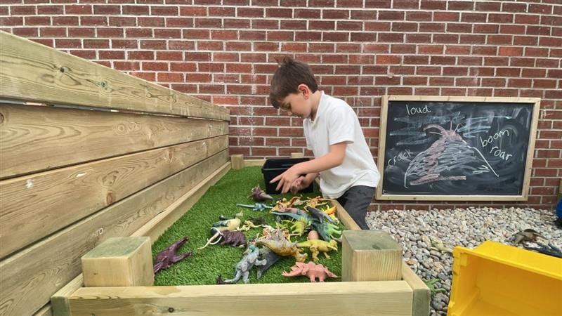 a little boy in black and white school uniform tips dinosaur toys onto the artificial grass surfaced construction table