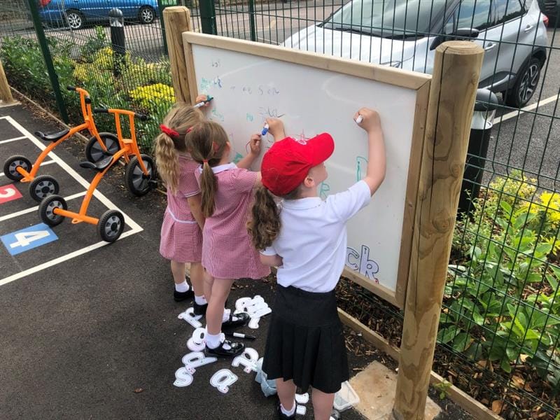 three children, in red, black and white school uniform write on the giant whiteboard in green and blue pens 