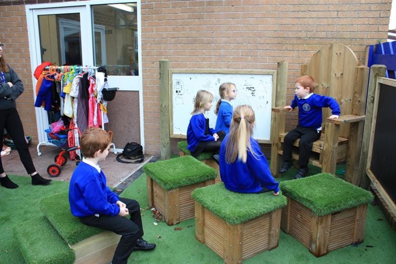 a little boy with auburn hair in a blue school jumper sits on the timber story-telling chair and talks to his classmates, also in royal blue school uniform. 