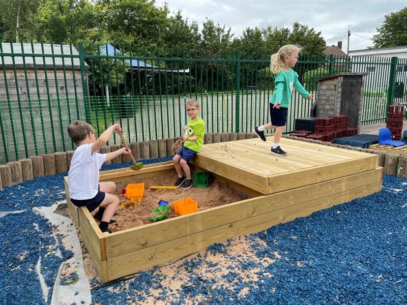 children in shorts and t-shirts run around in the sand pit with spades and buckets