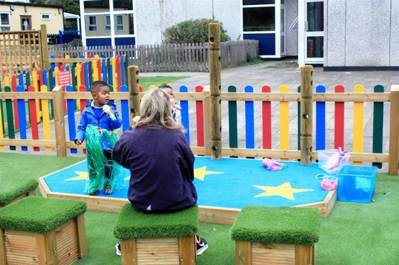 two little boys stand on the stage and perform for their teacher who sits on an individual grass topped seats