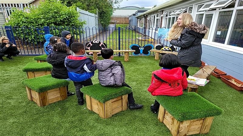 a teacher brings a story book to the story telling circle to read to the children sat on the grass seats