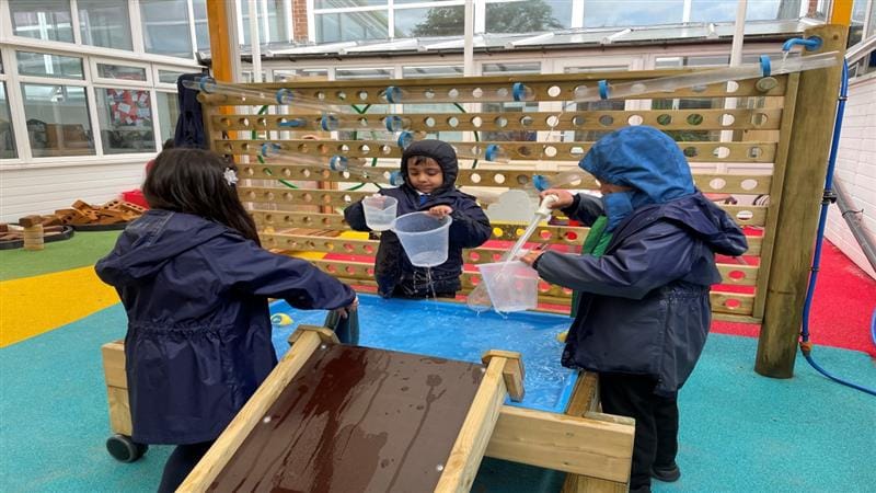 three children in winter coats gather around a damming station with a water wall in the background and pour water down