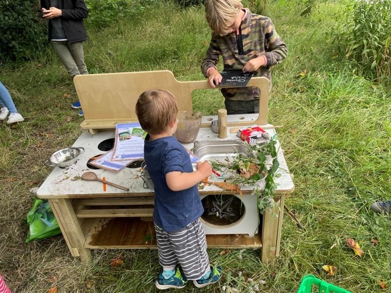 a little boy pretends to cook on the mud kitchen as an older child writes the menu specials