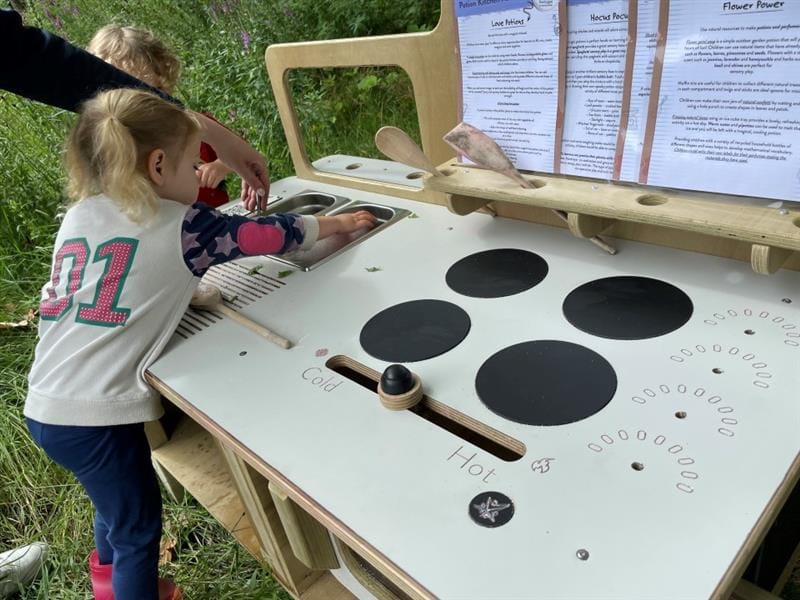 a little girl leans over the mud kitchen reaching into the sink as she reads the recipes propped on the menu holders