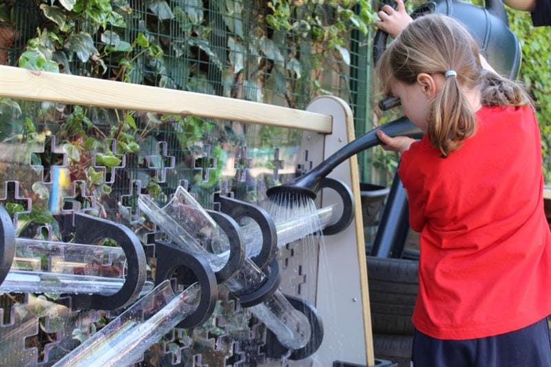 a child stands and pours water from a watering can down the channel and watches as it runs