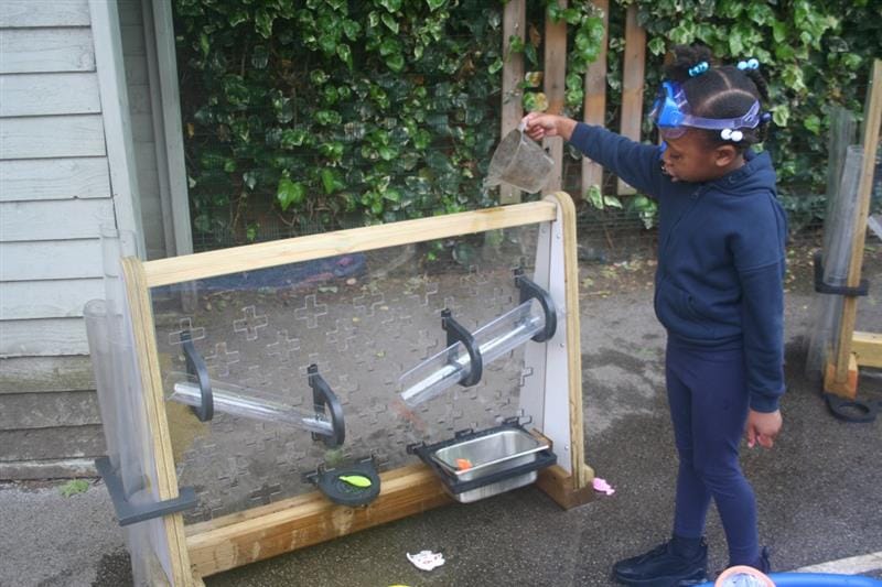 a little girl stands by the mini early years water wall and pours water down the channels using a measuring jug