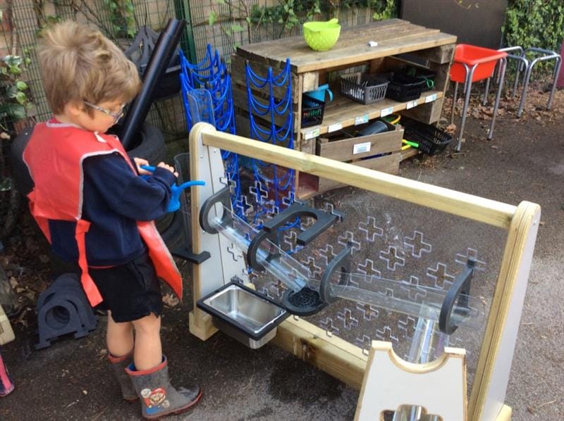 a little boy stands at the mini early years water wall and pours water down from the top