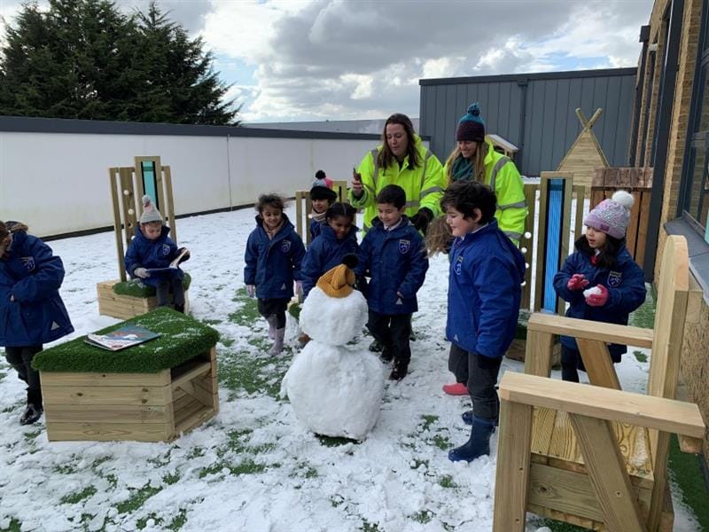 A group of children are playing in a play area which is covered in snow. Two adults are dressed in hi-vis and are watching over the children. A snowman has been built by the kids.