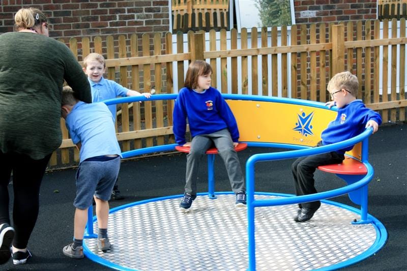 2 children sitting on a wheelchair roundabout seat whilst 2 children and an adult help push the roundabout