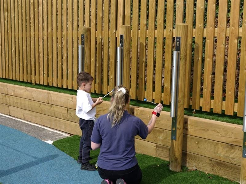 a child stands and hits the chimes with support from a teacher
