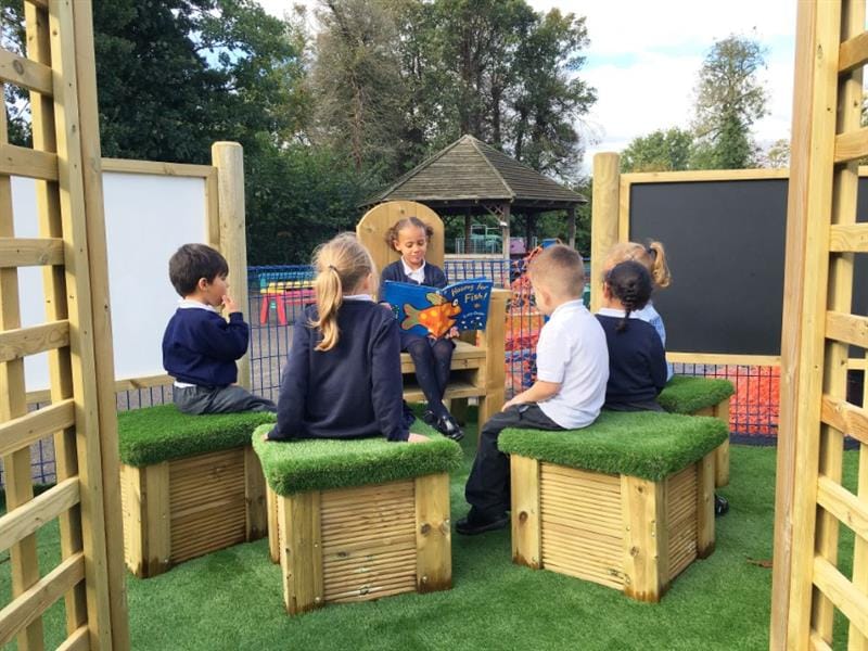 a child sitting on a storytelling chair reading out loud to 5 children sat on artificial grass topped seats