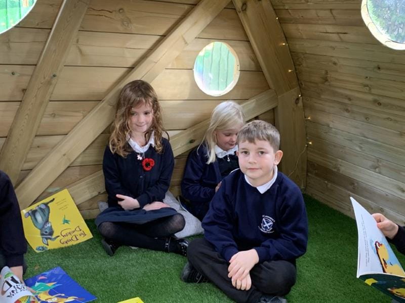 5 children sat on artificial grass inside of a hobbit house reading large books. Two girls wearing navy blue school summer dresses with one girl also wearing a red poppy, and one boy wearing a navy blue jumper and black trousers.