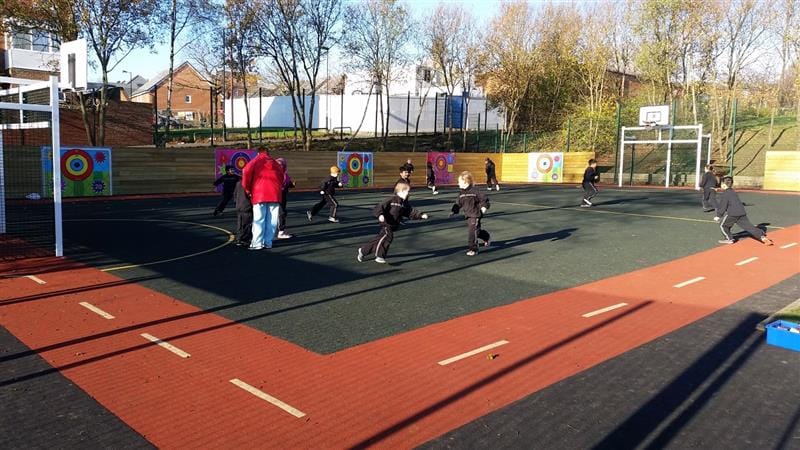11 children running around on school playground wearing black PE kits whilst one teacher wearing a red coat gives them instructions to follow.