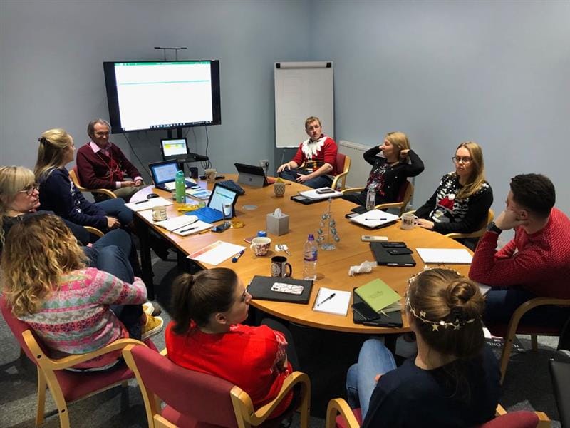 a team pentagon meeting gathered around a table with a whiteboard showing a presentation whilst dressed in Christmas attire
