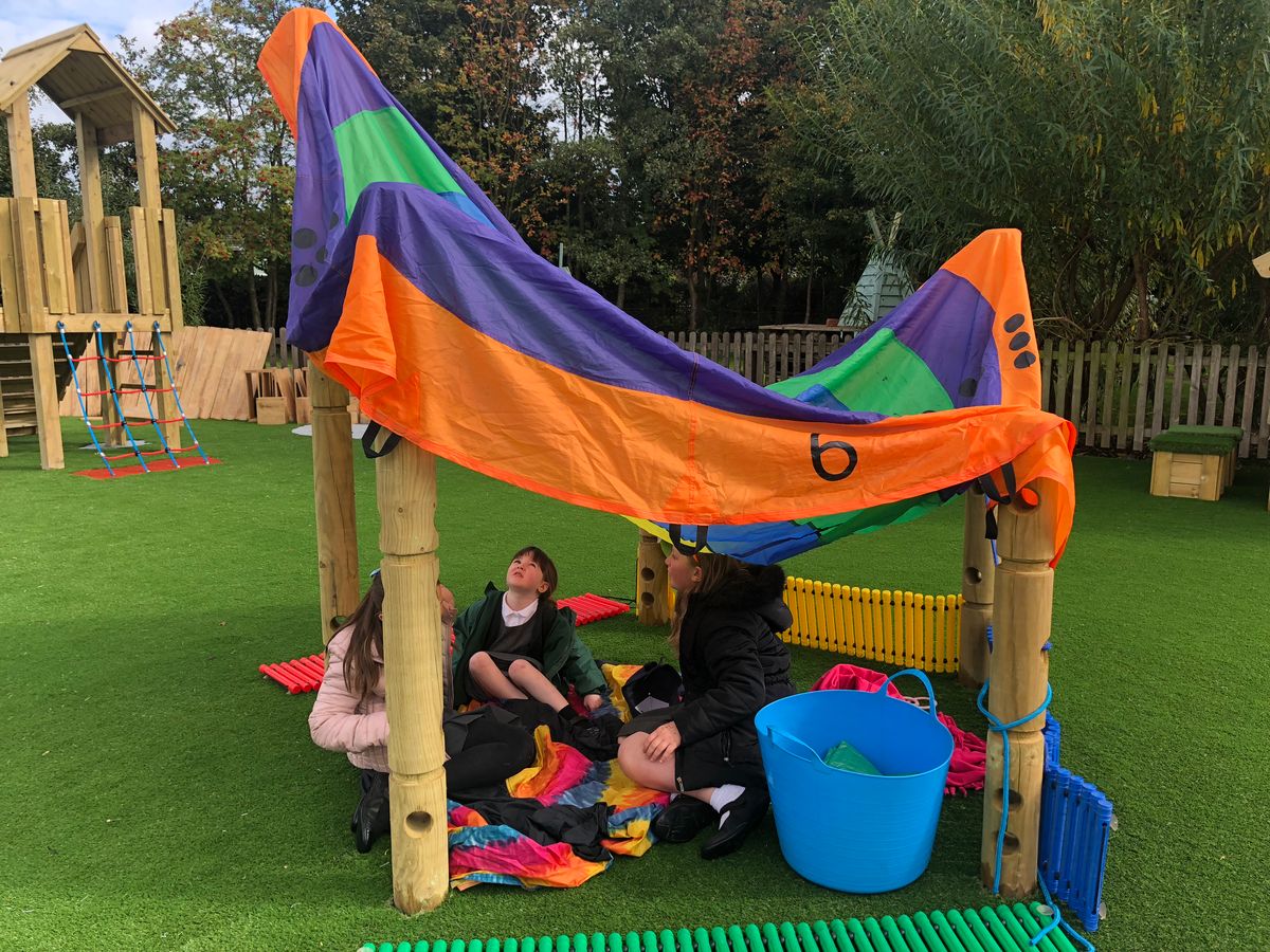 A small group of EYFS children are sat underneath a den that has been created by utilising a blanket and the Den Making Posts.
