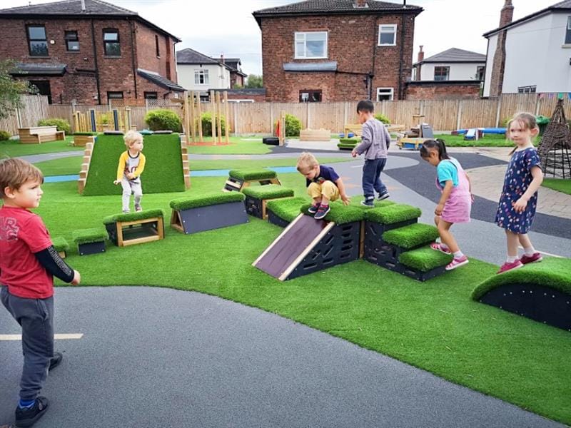 5 children playing on get set, go! blocks that have been placed onto artificial grass in the middle of a roadway whilst one boy wearing a red top with black sleeves stands on the roadway watching the children. 