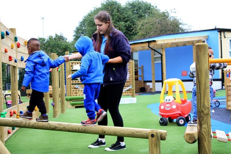 two children are helped along the balance beams by a teacher
