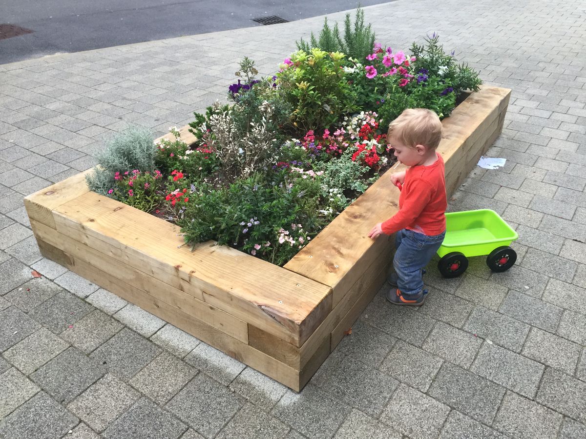 A little boy with a green pull cart has stopped to look at flowers that are growing from a planter.