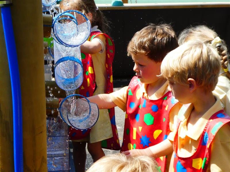 4 children wearing red waterproof bibs with multi-coloured shapes on them are playing with a water wall. There is water splashing out of the plastic tubes. 