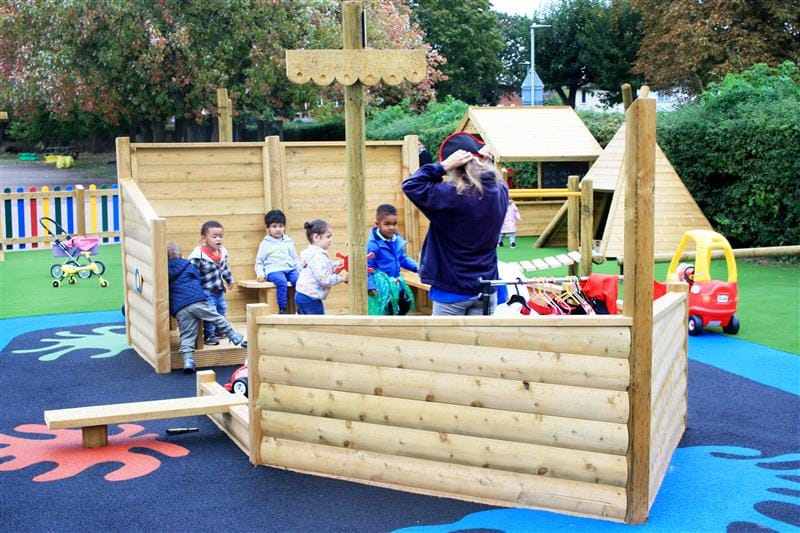 5 children playing inside of a pirate ship that has been installed onto blue splash wetpour surfacing whilst one teacher wearing a blue jacket and a pirate's hat stands at the front of the ship speaking to the children. 