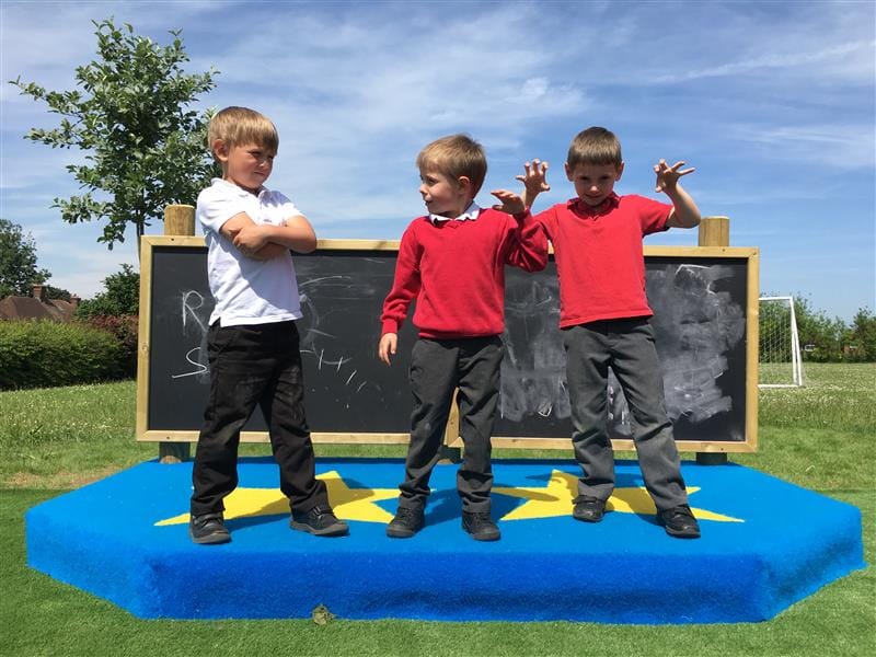 3 children stood on a performance stage, 2 wearing red school jumpers, one wearing a white t-shirt, they are stood in front of a chalkboard with scribbles on it. The performance stage has been installed onto the school field