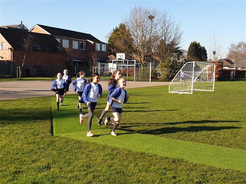 6 children, all girls wearing white polo tops and blue cardigans are running around the daily mile track which has been made using artificial grass and goes around the school field. 