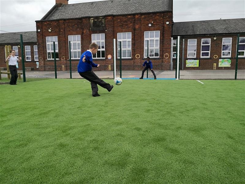 3 children playing on a MUGA pitch, one child wearing a blue jumper kicking a white and blue football into the football next whilst another child wearing a white top cheers at the side. 