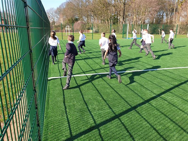 Close up photo of 13 children playing football on a MUGA pitch wearing white tops and navy blue jumpers with large trees behind the MUGA. 