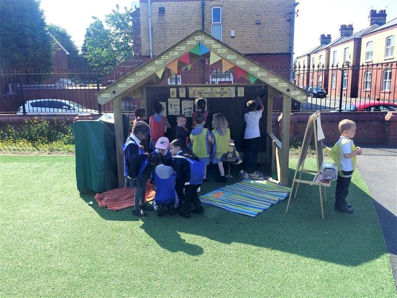 12 children wearing a mix of yellow, blue and red bibs are playing inside of a giant playhouse with a chalkboard that has been installed onto artificial grass in front of the school fencing. There are many houses on the other side of the fencing. 