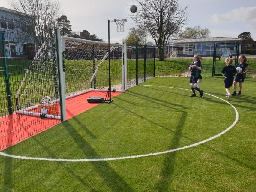 A small group of girls are throwing netballs towards a net, which is taking place on a MUGA.