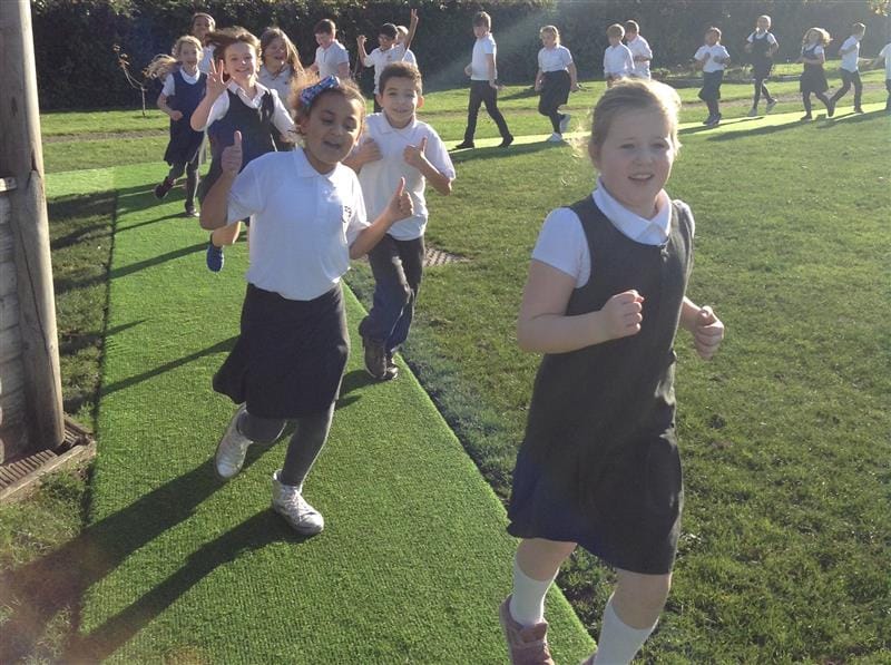 A class of children wearing white polo tops are running around a daily mile track which has been made out of artificial grass and installed onto the school field. Some children are smiling and doing thumbs up to the camera. 