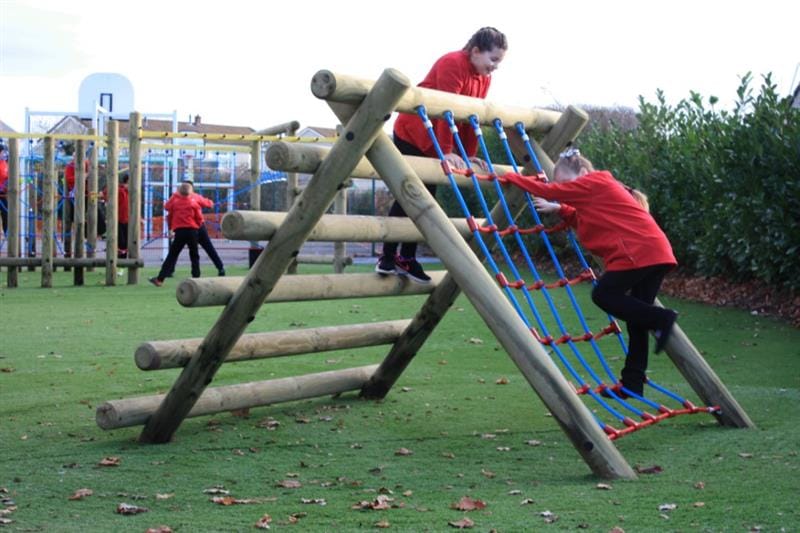 Two girls wearing red school jumpers are climbing over a rope climbing frame that has been installed onto artificial grass whilst more children play on a climbing frame in the background. 