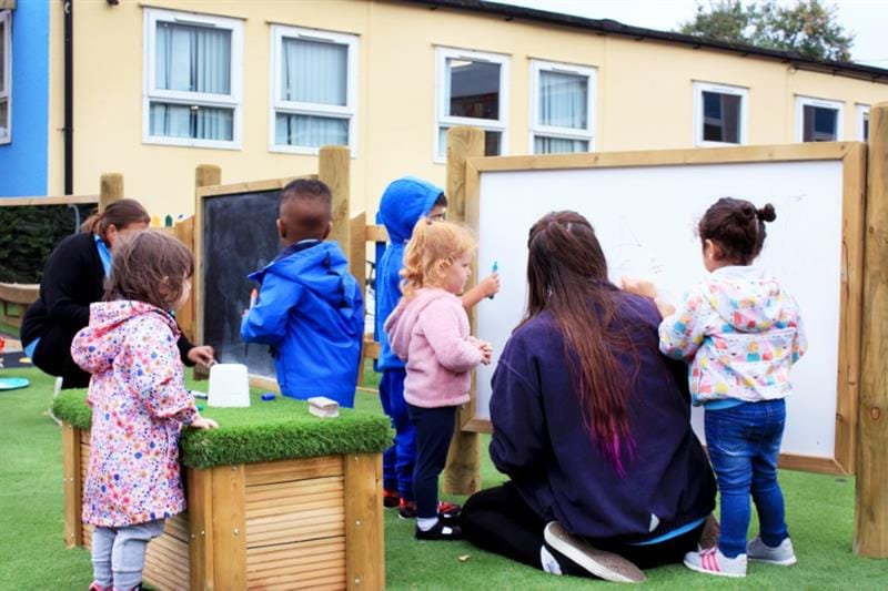 children kneel down by the whiteboard on posts with a teacher and draw on it