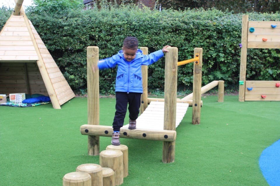 A little boy is crossing a wooden bridge as he takes a step onto the stepping logs.
