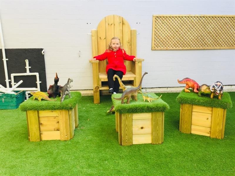 1 girl sat on storytelling chair wearing a red coat whilst toy dinosaurs sit on artificial grass topped seats in front of her. 