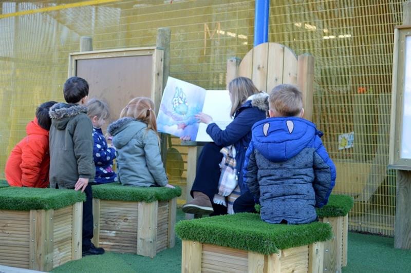 a teacher sits on the freestanding storytelling chair and reads a story book to the children sat on the grass topped seats