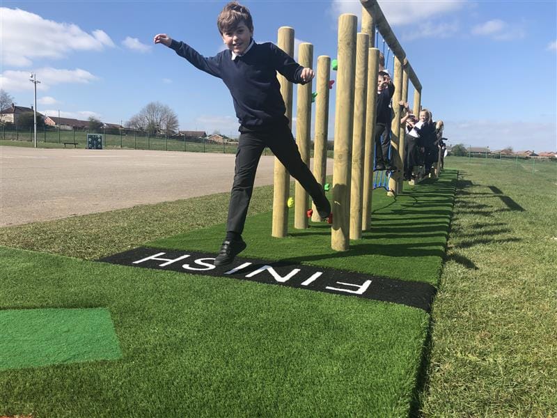 Children playing on a trim trail in the sunshine with one boy at the end of the challenge about to jump onto the black and white finish sign.
