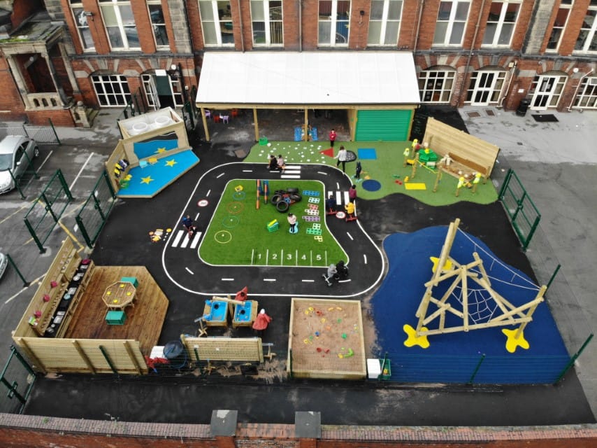 A tarmac playground with playground markings and areas of wetpour and artificial grass. On the playground, a variety of pieces of playground equipment can be seen, with children playing in all the different sections.