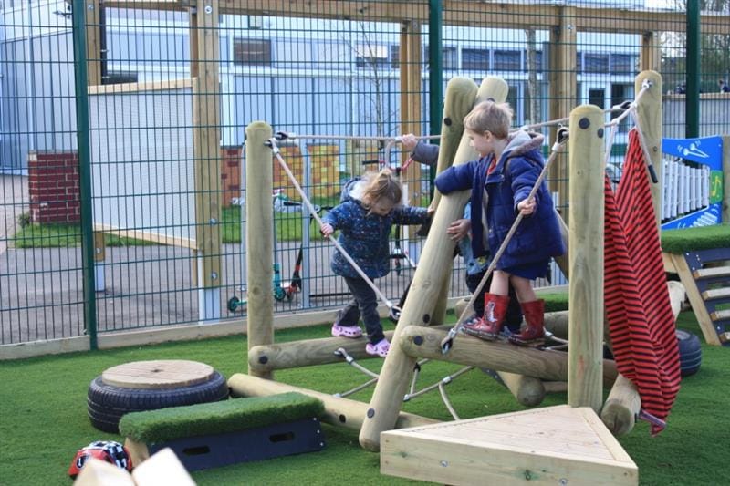 3 children playing on a small climbing frame that has been installed onto the artificial grass, one child wearing red wellington boots and one child wearing pink croc shoes. 