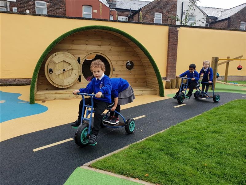 4 children playing on trikes on a roadway, two boys driving the trikes and 2 girls stood on the backs of the trikes all wearing blue school jumpers, installed in front of a hobit house.