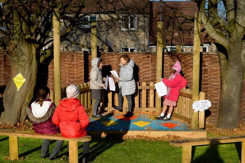 3 girls stood on a performance stage that has been installed in between two large trees, girls are reading a script from pieces of paper whilst 2 more children sit on the benches in the audience watching the show.  